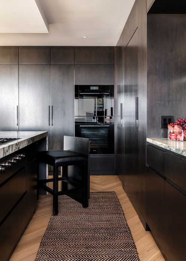 A refined galley-style kitchen with full-height dark timber cabinetry, integrated black appliances, and contrasting light stone benchtops. A central island with a black bar stool occupies the foreground, while a patterned runner sits over the light timber herringbone floor. The space has a clean, sophisticated, high-contrast contemporary look with recessed ceiling lighting and extensive built-in storage.