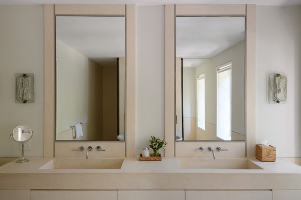 A bright, refined double-basin bathroom vanity with a long pale stone benchtop, two integrated rectangular sinks, wall-mounted chrome tapware, and twin large mirrors framed in light timber or painted joinery. Pale neutral walls, built-in cabinetry, small decorative wall sconces or artworks, and a floral arrangement create a calm, minimal contemporary interior. The mirrors reflect a hallway or adjacent space with windows.