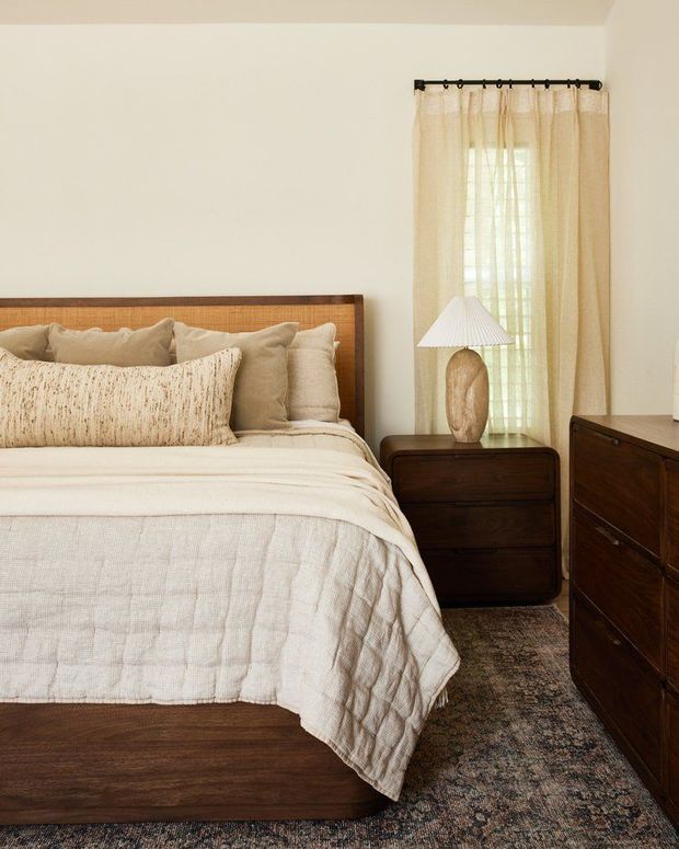 A warm, softly lit bedroom featuring a dark wood bed frame with woven cane headboard, layered cream and beige bedding, matching dark wood bedside table and dresser, a ceramic table lamp, sheer curtains, and a textured neutral rug.
