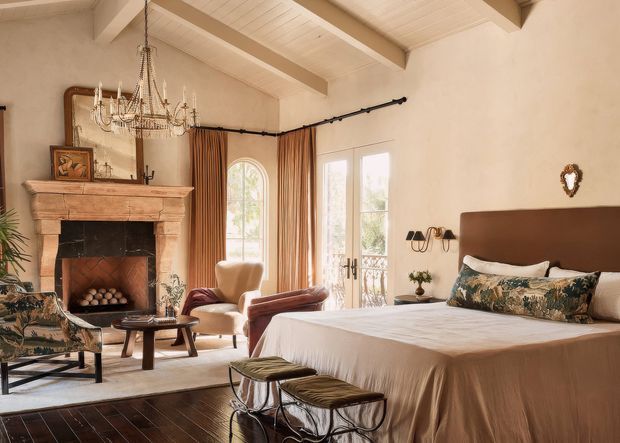 A warm, elegant bedroom with a high painted timber-lined ceiling and exposed beams. The room includes a large bed with a dark upholstered headboard, patterned cushions, two metal-framed benches at the foot, a carved stone or timber fireplace with a dark surround, an ornate chandelier, upholstered lounge chairs, tall arched and French doors, and full-height brown curtains. The styling blends traditional architectural details with refined vintage furnishings.