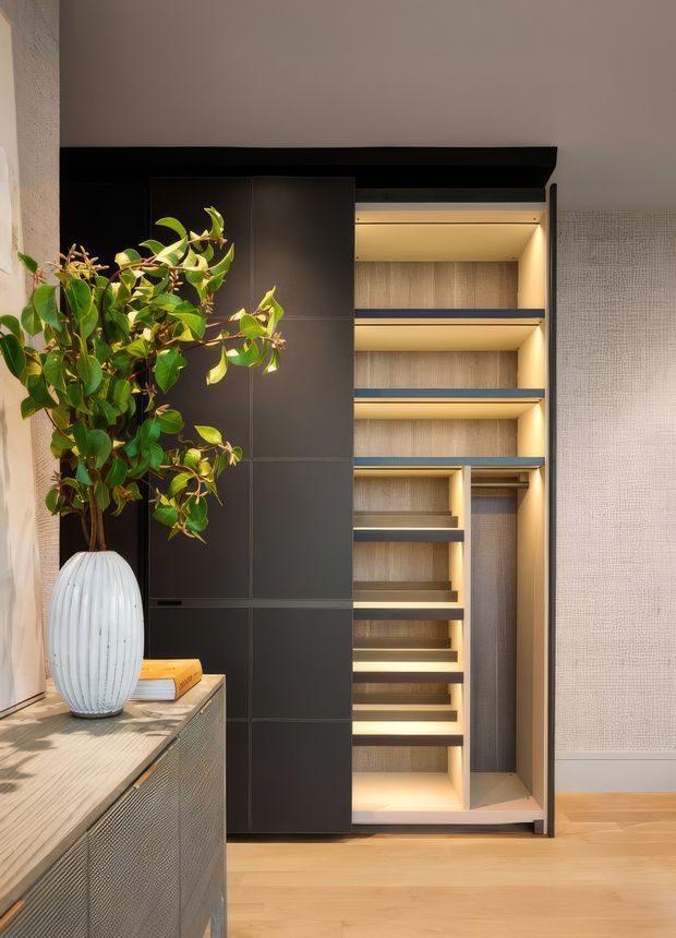 A warmly lit interior vignette focused on a tall, built-in dark cabinetry and open shelving unit with integrated LED lighting. The joinery includes multiple display shelves and a concealed or wardrobe-like section, set against textured neutral wall covering. In the foreground is a textured console with a pale wood-toned top, a white ribbed ceramic vase containing a leafy plant, and a small book. Light timber flooring and restrained contemporary detailing create a refined, softly contrasting composition.