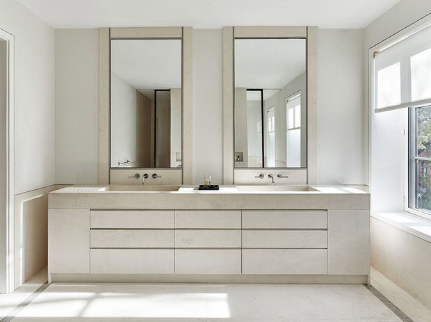 Bright, contemporary bathroom with a wide double vanity, two inset rectangular mirrors, wall-mounted chrome tapware, pale stone-look benchtop and matching light cabinetry. Large windows and a glazed door bring in abundant natural light, while the neutral finishes create a clean, refined appearance.