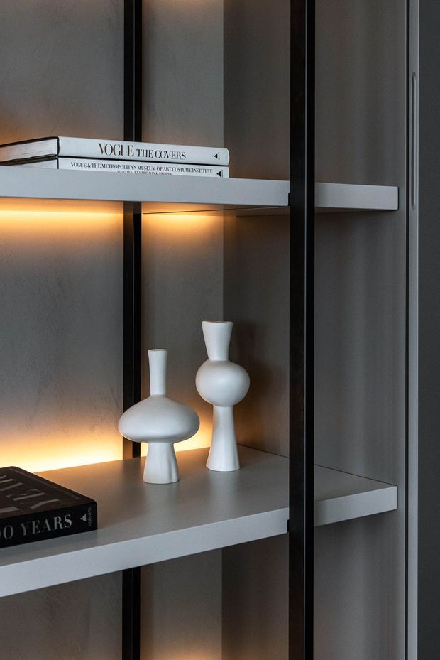 Close-up interior detail of a contemporary built-in display shelving unit. Matte grey shelves and a textured grey backing are framed by slim black vertical supports, with warm integrated LED strip lighting illuminating white sculptural vases and design books. A grey cabinetry or wall panel is visible at the right edge.