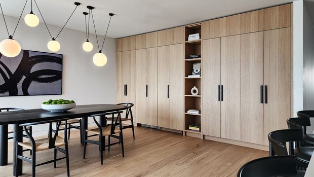 Contemporary dining area with a long dark dining table and black-framed chairs, beside a full-height wall of light timber built-in cabinetry with open display shelving. Multiple spherical pendant lights hang from a white ceiling, while an abstract monochrome artwork provides contrast. The space has a warm, refined, minimalist character.