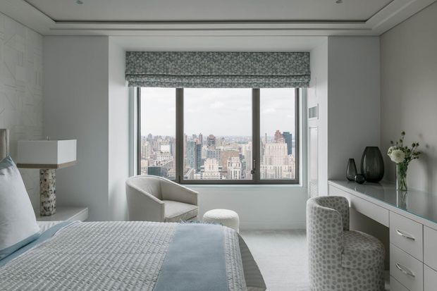 A refined, bright bedroom with a large picture window overlooking a dense city skyline. The room features a pale upholstered bed, textured light bedding, a curved accent chair and ottoman, a patterned upholstered desk chair, and a long white built-in dresser beneath a large mirror. Soft grey and white finishes, recessed ceiling detailing, and a patterned Roman blind create a calm contemporary interior.