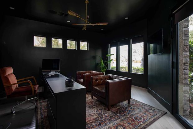 A dark, moody home office with black walls and ceiling, a long black built-in desk, computer workstation, leather office chair, and two brown leather lounge chairs arranged over a richly patterned area rug. Large windows and a glazed door bring in natural light, while a sculptural timber-and-brass ceiling fan adds a distinctive feature. A stone-clad exterior wall is visible at the right edge.