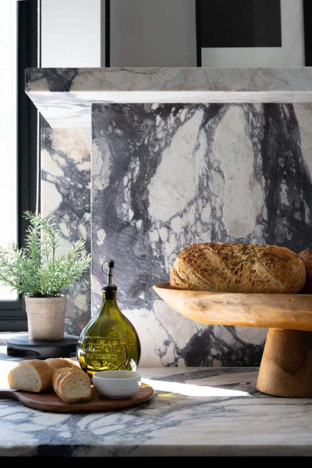 Close-up interior vignette featuring dramatic dark-veined marble or stone wall cladding and matching countertop, a sculptural timber pedestal table, bread and olive oil styling, and a potted plant beside a black-framed window. The image emphasises luxurious natural materials and strong daylight, with no clearly identifiable full room layout.