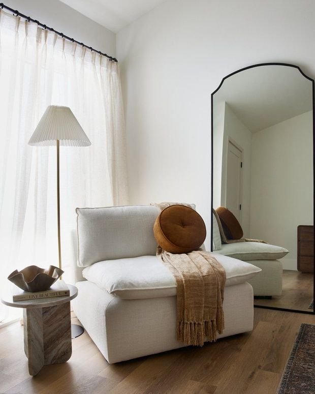 A softly lit, neutral-toned bedroom corner featuring a plush white upholstered armchair, a round rust-brown cushion, and a textured throw. Sheer curtains filter daylight beside a slim floor lamp, while a large organically shaped black-framed mirror reflects the chair and a doorway. The room has pale walls, warm timber flooring, a marble-look side table, and restrained contemporary decor.