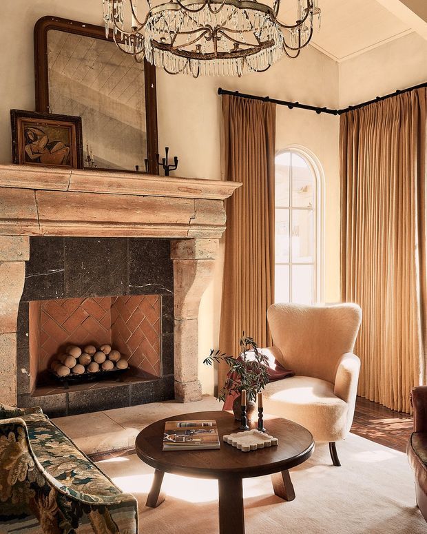Warm, richly layered living room featuring a large rustic stone and timber fireplace with herringbone brick firebox, an oversized antique-style mirror, ornate crystal chandelier, full-height pleated curtains, arched window, upholstered armchair, patterned sofa, and a dark round timber coffee table. The room blends traditional architectural detailing with refined, vintage-inspired furnishings and soft natural light.