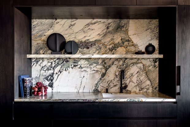 A dramatic, dark-toned kitchen or bar-style built-in joinery wall featuring a large veined natural stone splashback and benchtop, dark timber cabinetry, a slim open display shelf, black tapware, and minimal decorative objects. The composition is refined and luxurious with strong marble veining contrasted against deep wood tones.