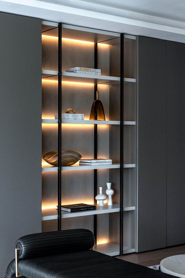 A close-up interior view of a sleek built-in display shelving unit with dark grey cabinetry, slim black vertical framing, illuminated open shelves, and curated decorative objects and books. A black ribbed lounge chair and part of a round light-coloured table are visible in the foreground. The space has a refined, moody contemporary feel with warm integrated lighting.