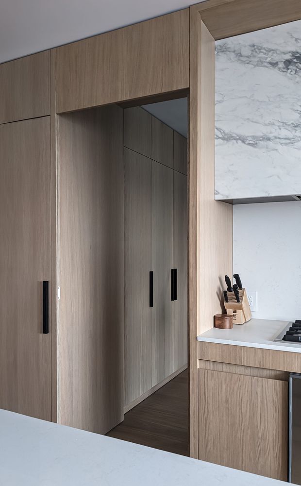 Close-up view of a refined contemporary kitchen with full-height warm wood cabinetry, a concealed passage or pantry door, pale stone benchtops, a marble-look splashback or wall panel, and black integrated hardware and cooktop. The image emphasizes the joinery and material palette rather than the entire room.