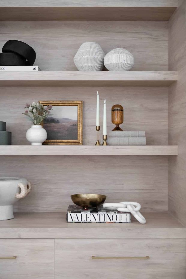Close-up of a built-in light timber display shelving unit with three open shelves and lower cabinetry. The shelves are styled with neutral ceramics, decorative objects, books, candles, a framed landscape artwork, and a brass-toned bowl. The joinery has a soft, pale wood finish with slim horizontal shelves and long brass handles, creating a refined, warm and curated interior vignette.