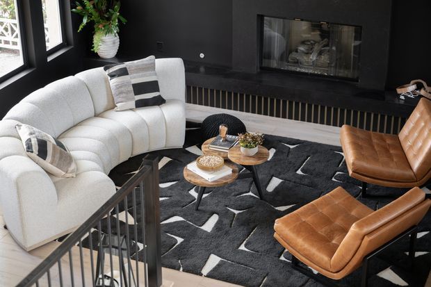 A stylish contemporary living room viewed from a stairway, featuring a large curved cream sectional sofa, tan leather lounge chairs, a black-and-white geometric rug, round wood coffee tables, and a dark fireplace wall with integrated firebox and vertical slatted detail. Large black-framed windows bring in natural light, while a potted plant adds greenery.