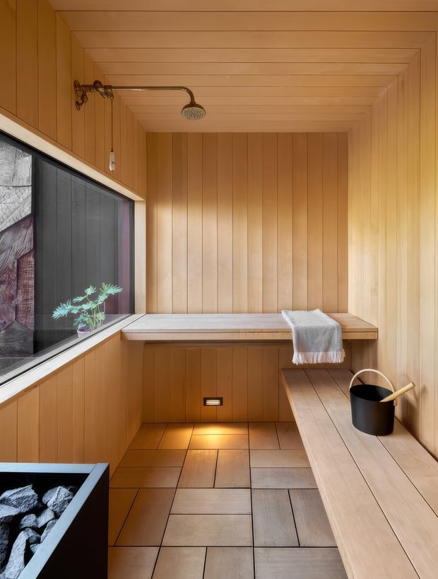 A compact wood-lined sauna with vertical timber wall panelling, a stepped timber ceiling, built-in corner bench seating, and a lower bench extending along the right wall. A small shower fixture is mounted near the ceiling, while a window brings in natural light. The floor is finished in large rectangular stone or ceramic tiles, and a black sauna bucket and ladle sit on the bench. A partial heated-stone stove is visible in the lower-left corner.