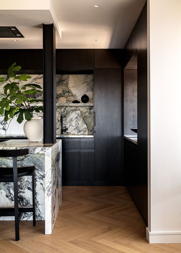 A sophisticated dark-toned kitchen interior viewed from an adjacent open area. Full-height black timber cabinetry frames a marble-lined cooking and display zone, with a dramatic veined marble island and integrated seating in the foreground. Warm herringbone timber flooring, black architectural columns, recessed ceiling lights, and a large leafy plant create a refined, high-contrast interior.