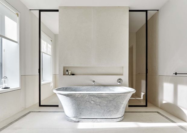 Bright, refined bathroom featuring a freestanding sculptural stone bathtub centred in front of a pale textured feature wall with a recessed shelf and wall-mounted tapware. Black-framed glazed shower or enclosure panels create strong linear contrast, while pale tiled surfaces, large windows and abundant daylight give the room an airy, minimalist character.
