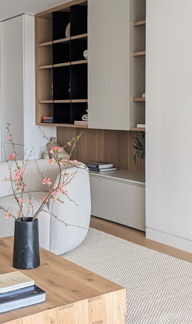 A softly styled living room with a built-in wall of pale cabinetry, open shelving, and a warm vertical timber slat feature behind a low media or storage unit. A light upholstered lounge chair, natural timber coffee table, textured neutral rug, books, and a black vase with flowering branches create a calm, refined interior.