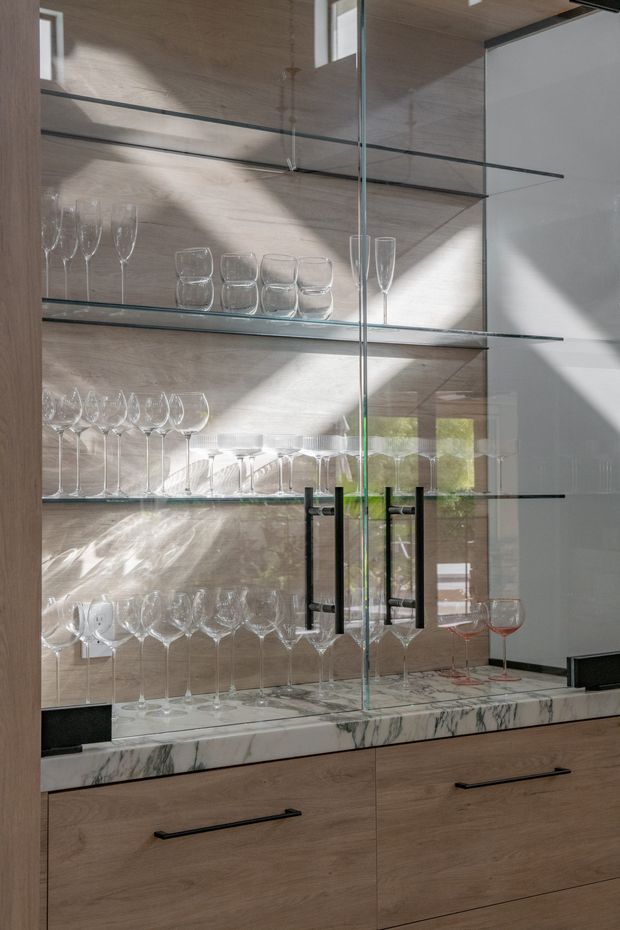 Close-up interior view of a bespoke glass-fronted drinks and wine cabinet. Full-height glass doors with black metal handles enclose multiple glass shelves displaying wine glasses and tumblers. The cabinet has light timber veneer panels, a dramatic white marble benchtop with grey veining, and dark trim, with sunlight creating strong reflections and shadows.