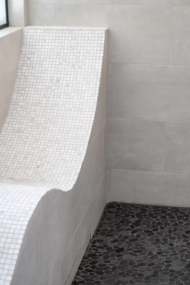 Close-up of a minimalist tiled wet-room or bathroom corner featuring a curved built-in bench or seating ledge finished in small white mosaic tiles. Large light-grey wall tiles surround the area, while the floor is covered in dark irregular natural pebble tiles. A small window is visible at the upper left.