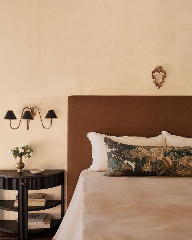 A warmly styled bedroom featuring a large upholstered brown headboard, cream bedding, and a decorative patterned lumbar pillow. A black fluted bedside console with open shelving, books, and a small floral arrangement sits beneath three dark wall sconces. The beige plaster wall includes a small ornate decorative mirror or wall accent, creating a refined, traditional setting with layered textures and warm neutral tones.
