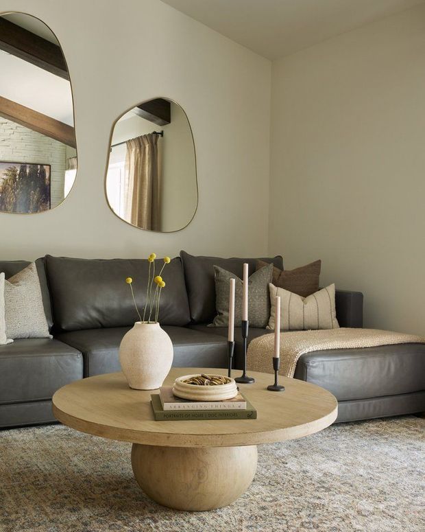 A warm, softly styled living room featuring a dark charcoal-grey sectional sofa, textured neutral cushions, a rounded light timber coffee table with a pedestal base, sculptural ceramic vase, taper candles, and a pale textured rug. The walls are painted a soft warm off-white, with two organic-shaped wall mirrors reflecting a brick feature wall, timber beam, and curtained window.