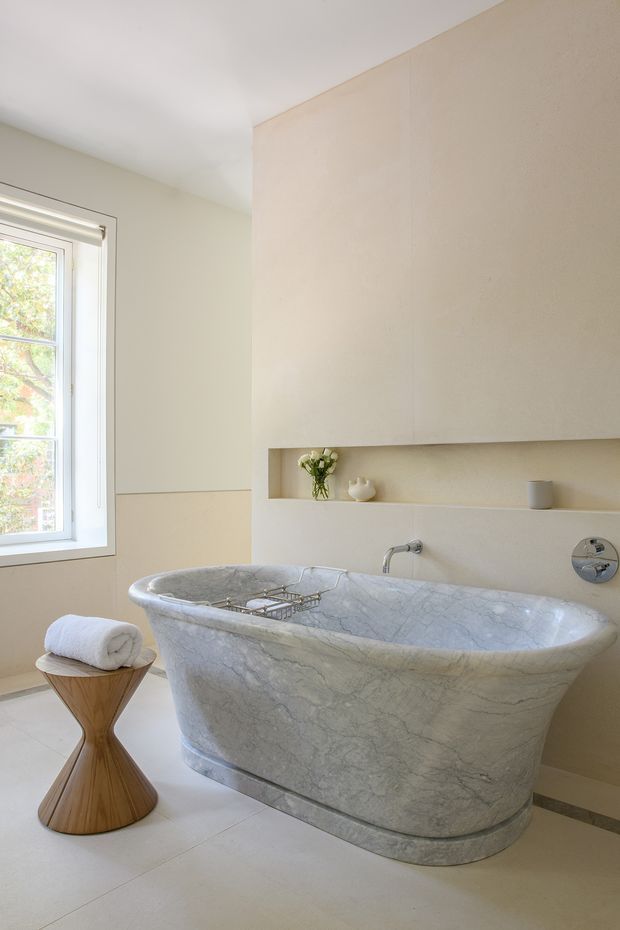 A bright, refined bathroom featuring a freestanding veined stone bathtub beneath a large flush wall cabinet with an open display niche. A wall-mounted chrome tap and controls sit above the tub, with a tall white-framed window providing natural light. Pale stone or tile flooring and warm neutral walls create a calm, luxurious atmosphere. A small sculptural wooden stool with a rolled towel is positioned beside the bath.