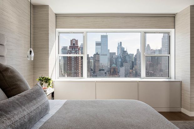 A contemporary bedroom with a large picture window overlooking a dense urban skyline. The room features a low upholstered bed in grey, pale wood-grain wall finishes, built-in cabinetry beneath the window, a slim pendant light, and a small indoor plant. The design is restrained, neutral, and space-efficient, with strong city views.