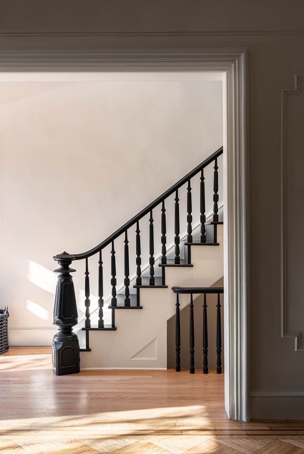 A refined interior view through a wide, heavily moulded doorway showing a traditional staircase with black turned balusters, a sculptural newel post, and dark handrail. The staircase rises along a pale plaster wall with decorative panel moulding, while warm timber flooring, layered door casing, and strong sunlight create a classic, elegant atmosphere.