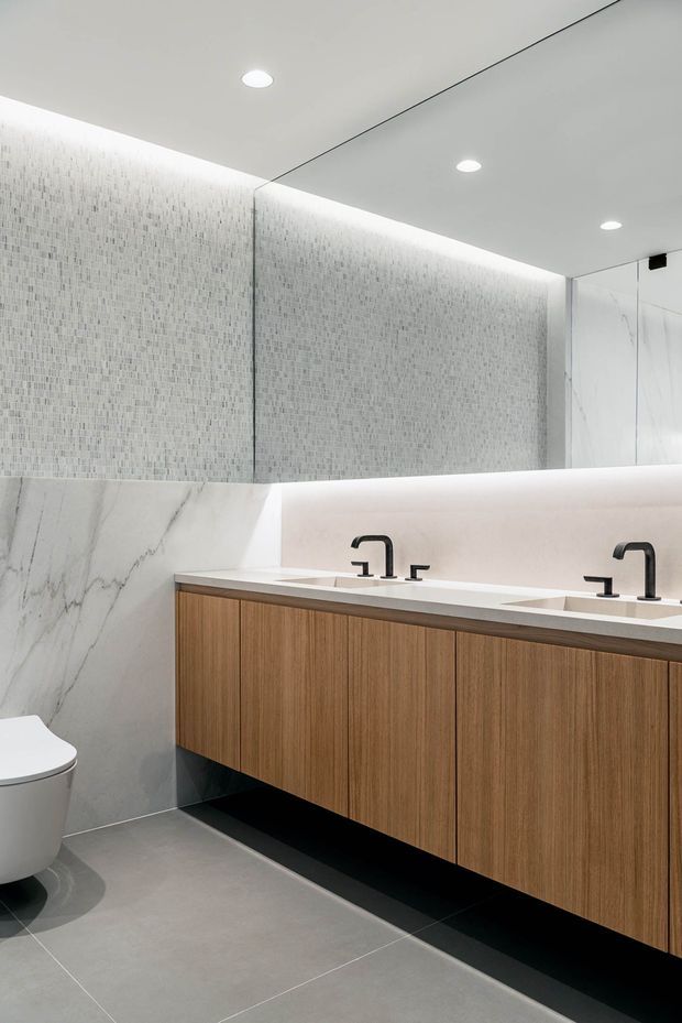 Contemporary bathroom with a long double vanity, warm timber cabinetry, white stone benchtop, black tapware, large illuminated mirror, and pale mosaic wall tiles. Large-format grey floor tiles and veined marble-look wall panels create a refined, minimalist finish.