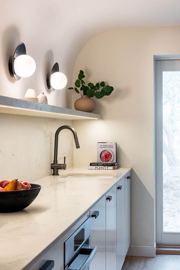A refined compact kitchen featuring pale blue-grey cabinetry, a light stone countertop and backsplash, matte black faucet and hardware, and a floating shelf with decorative objects and a potted plant. Two sculptural wall sconces illuminate the counter, while a glazed door or tall window provides daylight at right. The space has softly rounded wall detailing and warm wood flooring.