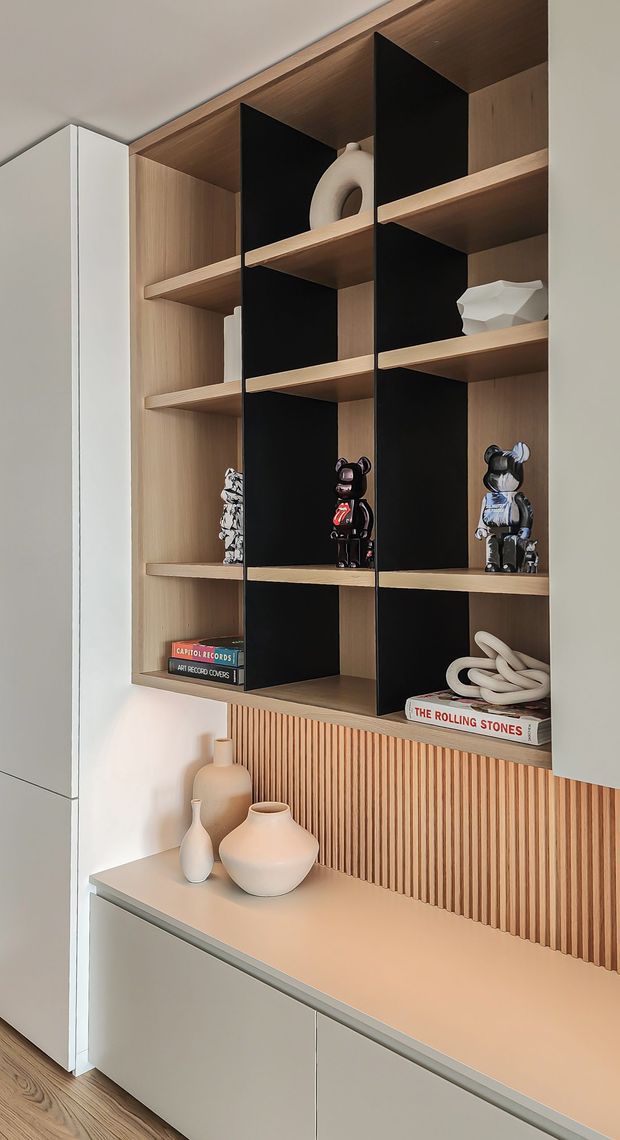 A close-up of a contemporary built-in storage and display wall. Warm timber shelving is divided by bold black vertical backing panels, with curated ceramics, art toys, books and decorative objects. Pale cabinetry and a light benchtop sit below, backed by a vertical timber slat feature panel. The scheme is restrained and modern with warm wood and neutral finishes.