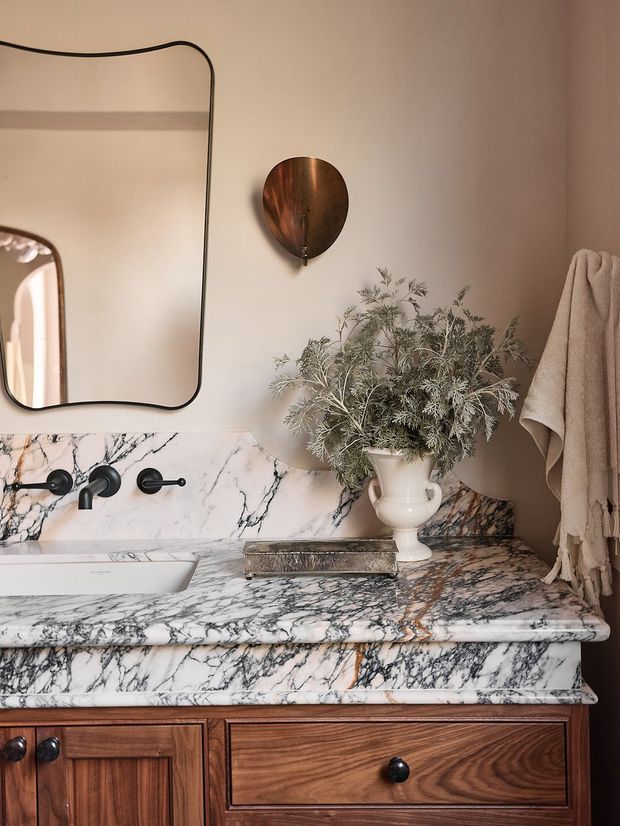 Close-up view of an elegant bathroom vanity featuring richly veined white marble across the countertop, backsplash, and basin surround, set above warm dark timber cabinetry. A rounded-edge wall mirror, vintage-style wall sconce, bronze-toned fixtures, neutral towels, and a white ceramic vase with foliage create a warm, refined look.