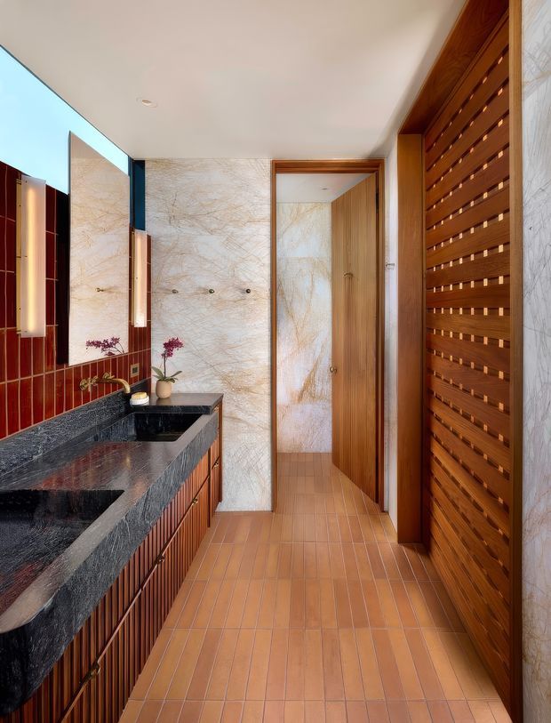 A richly detailed bathroom with a long dark stone vanity and integrated basins, warm fluted timber cabinetry, terracotta-toned tiled flooring, pale veined stone wall surfaces, and a striking red glazed tile feature wall. Timber slatted screening and matching timber doors add texture, while a large mirror and warm vertical wall lights enhance the space.