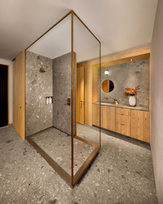 Contemporary bathroom featuring a large frameless glass shower enclosure with slim bronze-toned framing, terrazzo-style concrete flooring and wall surfaces, and warm timber cabinetry and doors. A long floating vanity with integrated stone-like countertop spans the rear wall, complemented by a round mirror, recessed lighting, and minimalist decorative styling.