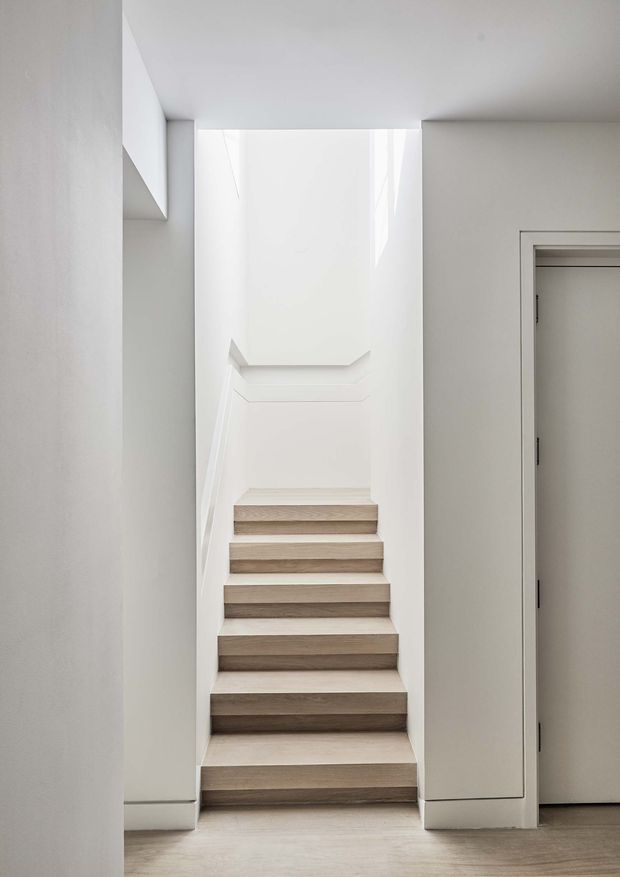 A minimalist interior stairway viewed straight on, with pale timber or wood-finished treads rising through a narrow white-painted stairwell. Clean flush walls, a simple integrated handrail, and a plain doorway on the right create a bright, restrained architectural composition.