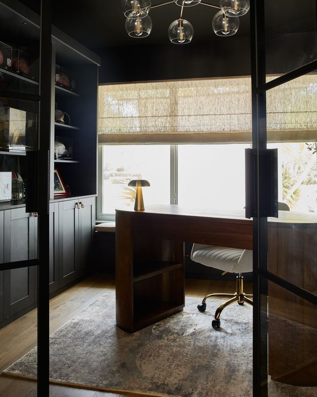 A compact, sophisticated home office enclosed by black-framed glass doors. The room features dark built-in cabinetry and shelving, a dark timber desk with integrated storage, a white upholstered office chair, a textured area rug, a sculptural desk lamp, and layered globe pendant lighting. Woven roller blinds filter daylight through the large window, creating a moody yet refined workspace.