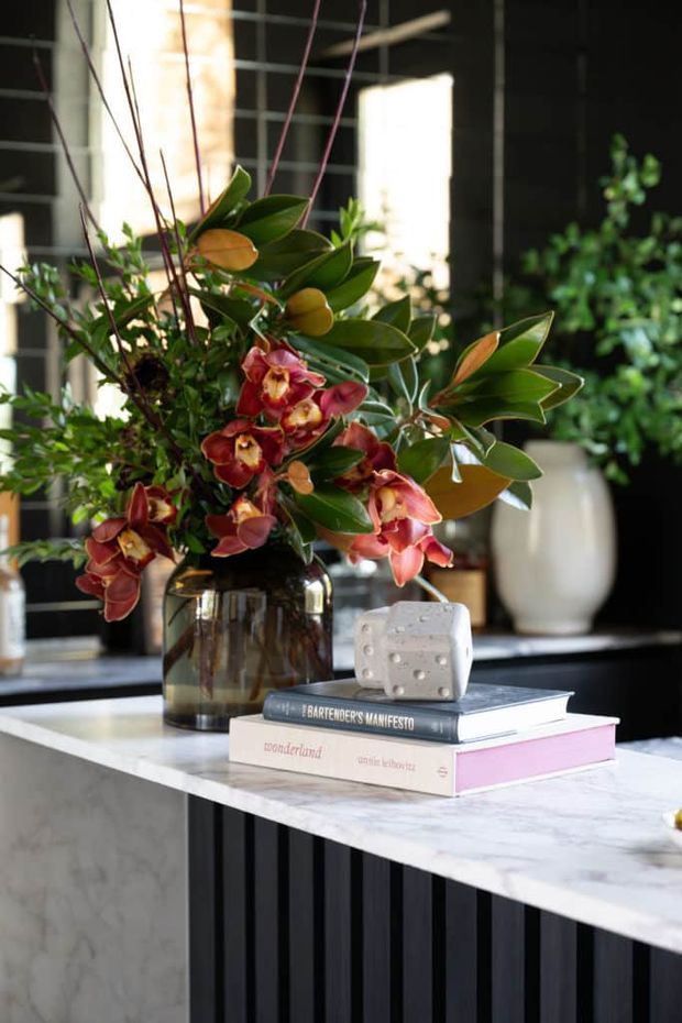 A styled close-up of a dark, sophisticated kitchen or bar counter, featuring a white marble-look benchtop with a black vertical fluted base. A smoky glass vase filled with a large arrangement of tropical greenery and burgundy flowers sits beside stacked design books and a small sculptural object. Black-framed glazing and additional greenery appear in the background.