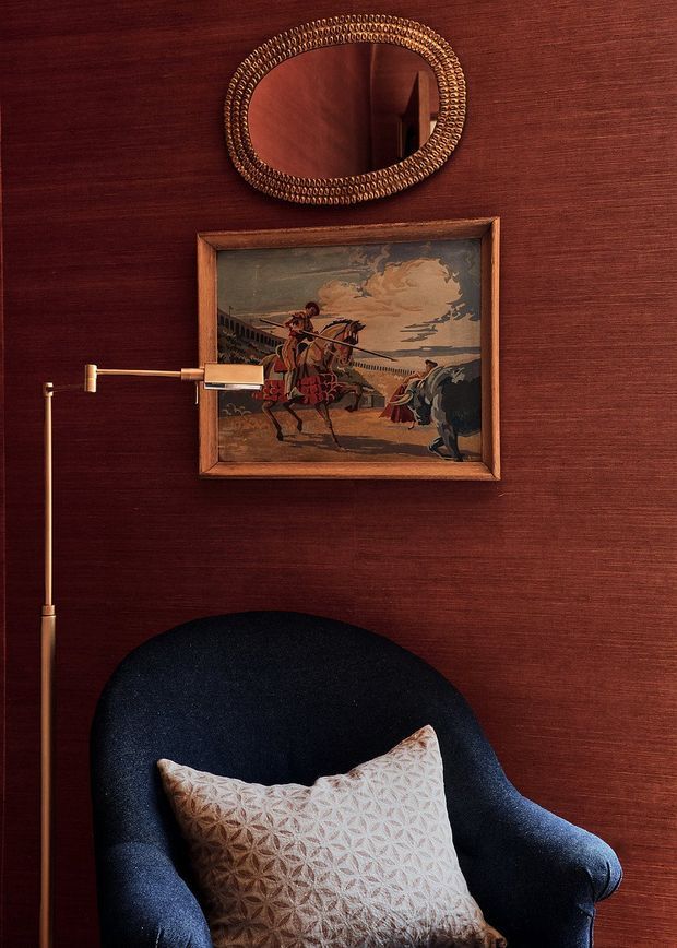 A richly styled reading nook with a deep navy upholstered armchair against a dark reddish-brown textured wall. A brass floor or wall-mounted swing-arm lamp extends beside the chair, while a patterned neutral cushion rests on the seat. Above are an oval decorative mirror with a braided metallic frame and a framed vintage equestrian artwork. The intimate composition has warm, moody, traditional detailing with strong burgundy, navy, brass, and timber tones.