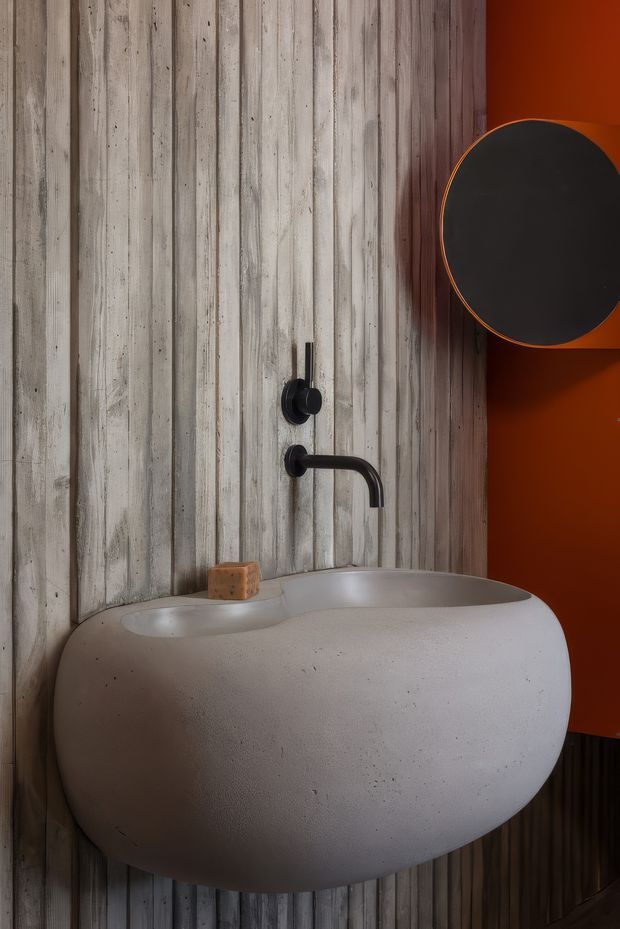Close-up view of a compact, dramatically styled bathroom featuring a rounded wall-mounted concrete or stone basin, matte black wall-mounted tapware, a vertically ribbed weathered timber wall finish, and a bold orange-red adjacent wall. A round black mirror with an orange edge is partially visible above the basin.
