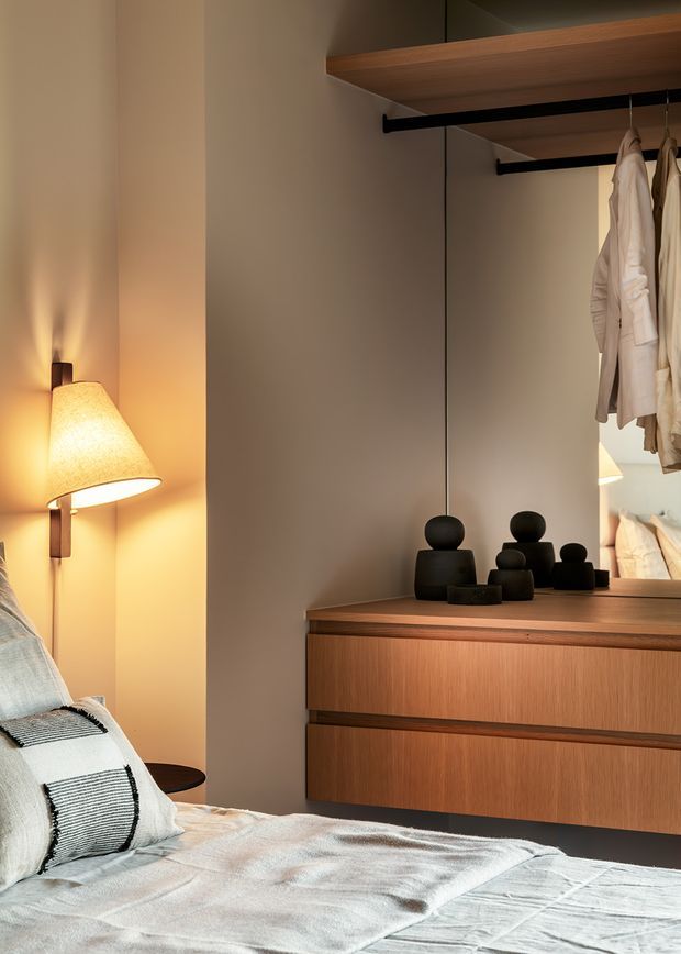 Warmly lit bedroom with a partially visible bed in the foreground, a wall-mounted bedside lamp, and a built-in timber wardrobe and dresser. The wardrobe has open hanging rails with clothing, dark decorative objects on the dresser, and mirrored or reflective panels that extend the view of the room. The design is understated and contemporary, with warm timber, taupe walls, black hardware, and soft neutral bedding.