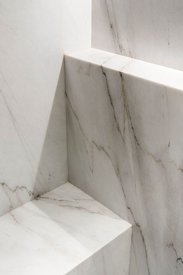 Close-up architectural detail of a minimalist stair or stepped built-in structure clad entirely in white veined natural stone. The image focuses on sharply folded planes, junctions, and a thick stone edge, with soft neutral lighting and no visible furnishings or wider room context.