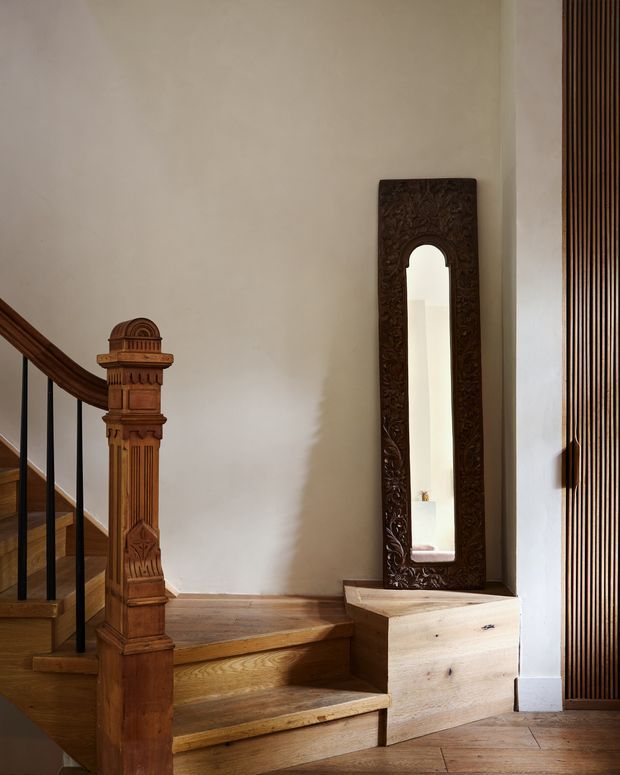 A warm residential stairway and landing featuring richly carved timber newel posts and handrail, timber stair treads and flooring, a tall ornate dark wood-framed mirror, and a vertical timber slat screen along the right wall. The space has a refined traditional character with natural wood detailing and soft neutral plaster walls.