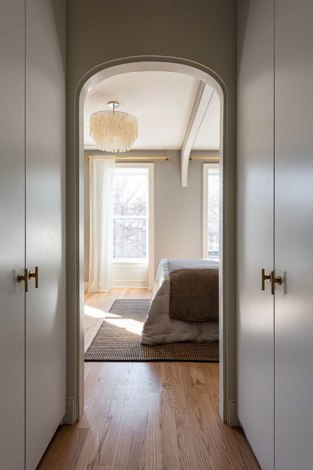 View through an arched doorway from a built-in wardrobe or hallway into a softly styled bedroom. The bedroom has light timber flooring, pale grey walls, tall windows with sheer curtains, a neutral upholstered bed, layered rugs, a decorative fringed pendant light, and a curved ceiling beam or architectural detail. White built-in cupboard doors with brass hardware frame the foreground.