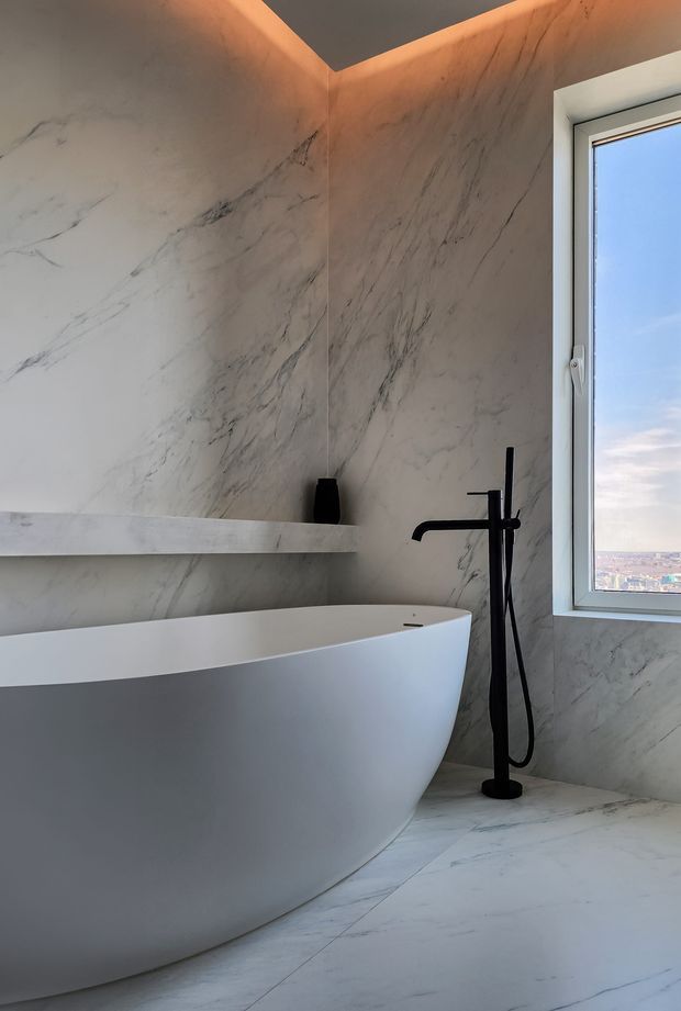 A refined contemporary bathroom featuring a sculptural freestanding white bathtub, full-height white marble-look wall and floor surfaces with grey veining, a slim floating stone ledge, and a matte black floor-mounted bath filler with hand shower. A tall window provides daylight and an expansive urban view, while warm concealed lighting highlights the ceiling edge.
