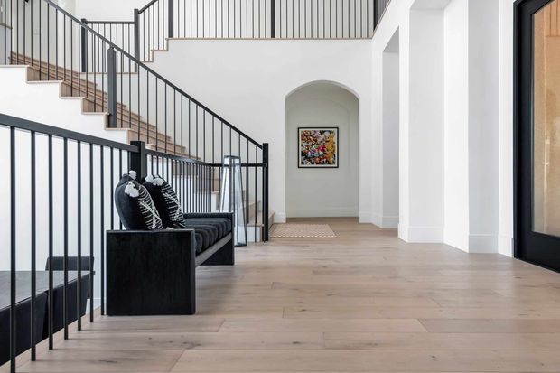 Bright contemporary interior hallway and stairway with pale timber flooring, crisp white walls, black metal balustrades, and a black built-in bench with patterned cushions. An arched recessed niche frames artwork at the end of the corridor, while a dark-framed glazed doorway is visible on the right.