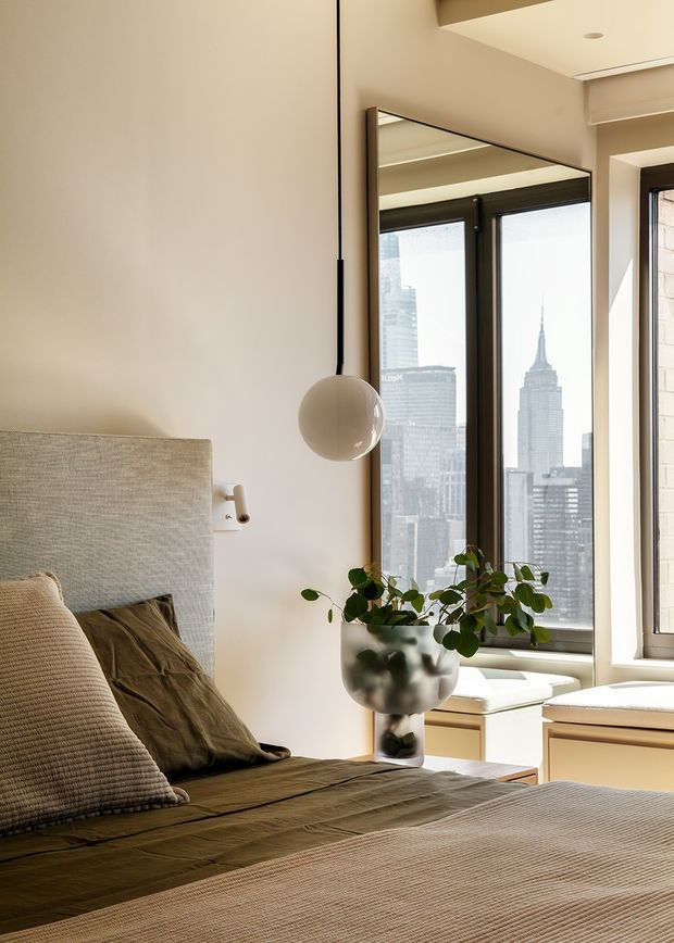 A warm, refined bedroom interior with a large upholstered bed, layered neutral and olive bedding, a spherical pendant light, and a sculptural potted plant beside tall black-framed windows. The windows reveal a dense city skyline, including the Empire State Building. Built-in window seating or cabinetry runs beneath the glazing, and a large mirror reflects the room and windows.