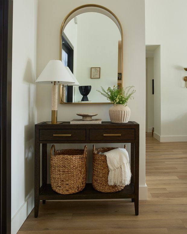 A refined entryway vignette featuring a dark timber console with brass drawer pulls, woven storage baskets, decorative objects, and a tall arched brass-framed mirror. The console sits against warm off-white walls beside a light timber floor, with a hallway and adjoining interior visible beyond. The styling is warm, understated, and contemporary with traditional accents.