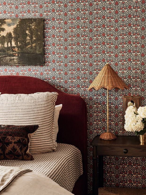 A richly layered bedroom corner featuring dark patterned wallpaper with a geometric floral motif, a deep burgundy upholstered bed, striped bedding and cushions, a dark timber bedside table, a woven conical table lamp, framed artwork, and a vase of white flowers. The room has a warm, dramatic, eclectic character with strong pattern and texture.