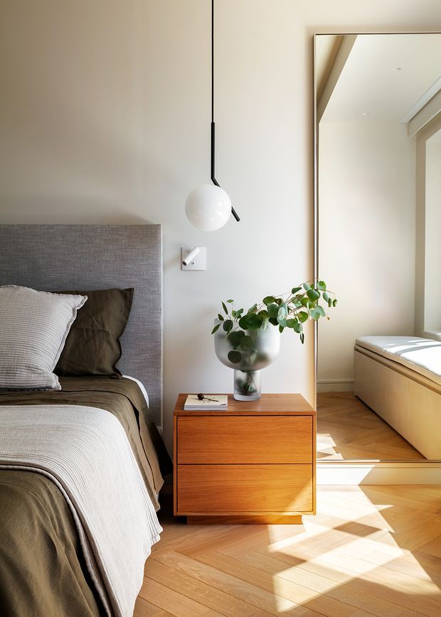A warmly lit bedroom corner with a low upholstered grey headboard, layered neutral bedding, and a walnut-toned two-drawer bedside cabinet. A spherical pendant light hangs beside the bed, while a potted leafy plant sits in a metallic vessel. A large framed mirror reflects a bright window seat and timber flooring, emphasizing the natural light and herringbone-style wood floor.